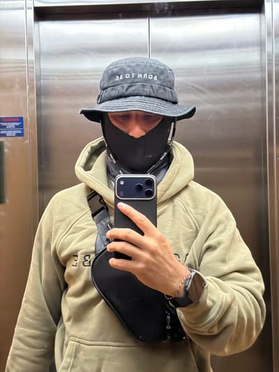 A person is standing in an elevator, holding a smartphone to take a mirror selfie. The person is wearing a dark bucket hat with white lettering on it, a black face mask, a light green hoodie, and a black crossbody bag. The elevator walls appear to be made of brushed metal. There is a blue and white sticker on the left elevator door, possibly an emergency notice. The person's right wrist is adorned with a thin bracelet and a smartwatch. The image does not provide any specific visual cues to indicate the location within Banyuwangi, Indonesia.