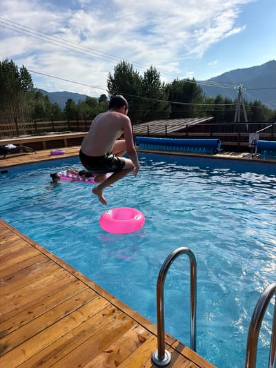 In Elekmonar, Russia, a teenage male is captured mid-air as he jumps into a bright blue swimming pool. He is wearing dark swim trunks and appears to be focused on his dive. A bright pink inflatable ring floats in the water below him. In the background, another person is visible in the pool, lying on a pink inflatable mattress. The pool is surrounded by a wooden deck. The environment is natural, with tall green trees and mountains visible in the distance under a partly cloudy sky. Electrical wires and poles are also present in the background, indicating the presence of infrastructure. The overall scene depicts a recreational summer day.