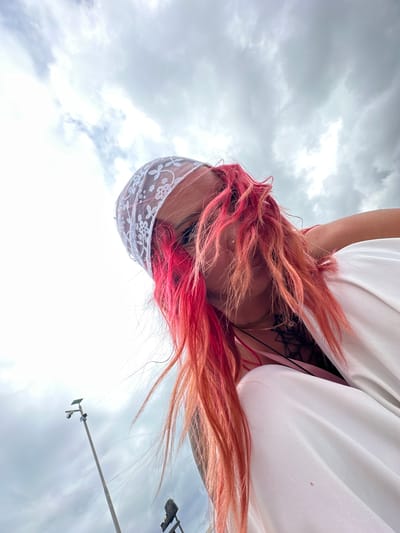 A low-angle shot depicts a person with vibrant pink and red hair wearing a white lace head covering. The person is looking down, and a white garment obscures much of the view. A black tattoo is visible on the upper chest. The backdrop features a cloudy sky and a lamppost. The location is Kampung Toyapakeh, Indonesia.