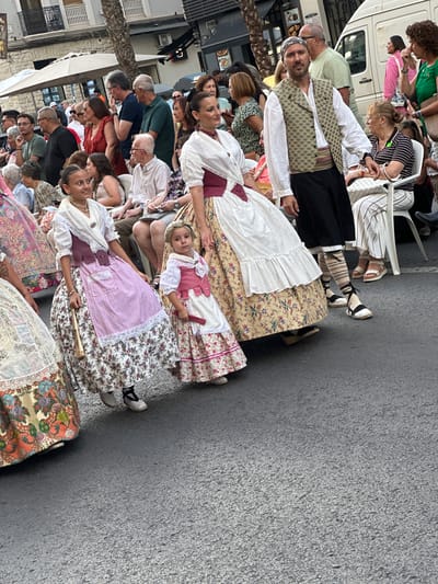 This image captures a street scene in Alicante, Spain, likely during a festival or parade. Several individuals are visible, dressed in traditional attire. In the foreground, two women and a young girl are wearing elaborate, full-length dresses with layered skirts and fitted bodices. The dresses feature intricate floral patterns, lace trim, and contrasting colors such as pink, white, and deep red. The women are adorned with white shawls or head coverings.

To the left, a portion of another woman's dress is visible, also ornate and colorful. Behind these figures, a man in a white shirt, patterned vest, and black pants stands with a serious expression. He wears a patterned headscarf.

In the middle ground, a crowd of onlookers is seated and standing, many also in festive or casual clothing. The backdrop includes buildings with balconies and windows, suggestive of urban architecture. A white van and a large umbrella are also present. The ground is paved with asphalt, and the lighting indicates it is daytime. The overall atmosphere suggests a cultural event taking place outdoors.