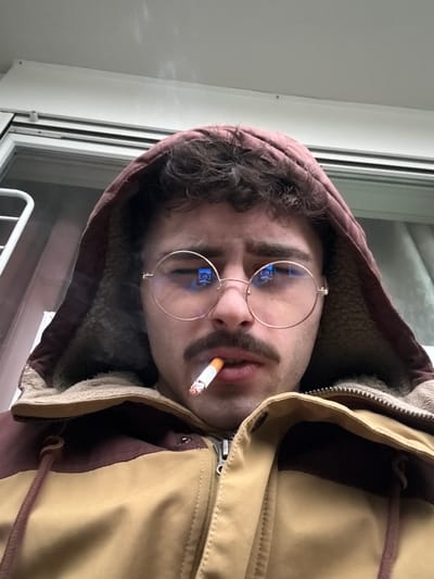 A man with fair skin, a dark mustache, and curly hair wears round glasses and a brown hooded jacket with a tan shoulder area. He holds a lit cigarette between his lips, smoke visible around it. Part of a white window frame is visible in the background, near the lower left. The image is slightly rotated clockwise.