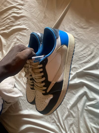 The image captures a close-up view of a pair of Nike Air Jordan 1 Low sneakers, held by a dark-skinned hand, resting on a crumpled, light-colored sheet, likely a bed, in Lakka, Sierra Leone. The sneakers display a distinct colorway, primarily white, black, and royal blue.

The shoe prominently features a white perforated toe box, white mid-panel, and a white portion on the heel. These white sections are contrasted with black leather on the toe cap, eyelet panel, and the rear quarter. A bright royal blue color is visible on the heel collar and the inner lining of the shoe. A standout detail is the cream-colored reverse Nike Swoosh on the lateral side, positioned against the white mid-panel. The laces are also cream-colored, and the midsole is an off-white hue. Visible branding includes a blue "NIKE AIR" text on the tongue tag and a partial white "Wings" logo on the black heel tab of the held shoe.

The hand, with its dark skin tone, is holding one shoe upright, with the second shoe of the pair partially visible underneath. The activity appears to be showcasing or inspecting the footwear. The setting is an indoor space, likely a bedroom, with natural light entering from the left, indicated by the shadows cast on the wrinkled sheet. No specific time of day beyond daytime is discernible, and no emotions are conveyed.
