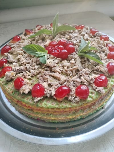 A circular, multi-layered cake is presented on a silver platter. The cake has distinct layers of what appear to be colorful crepes or thin pancakes, in shades of green, yellow, and red. The top of the cake is adorned with a crumbly topping, possibly crushed cookies or nuts, interspersed with bright red maraschino cherries. Several fresh mint leaves are strategically placed on top as a garnish. The cake is positioned on a table with a white lace tablecloth and a light-colored wall in the background. The image does not provide any specific details to locate it within the city of Lykhove, Ukraine.