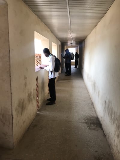 The image displays a long, narrow indoor hallway or corridor, viewed lengthwise, creating a strong perspective toward a brighter area at its far end. The floor and ceiling are made of concrete. The left wall (from the viewer's perspective, before rotation) is clad with vertical, light-colored paneling, featuring a white electrical conduit running horizontally along it. The right wall is textured concrete with a speckled finish. On this wall are multiple rectangular window openings, one of which in the foreground has a reddish-brown decorative grate.

Three young men are present in the hallway. The closest individual, positioned in the right foreground, stands looking towards the grated window. He wears a white long-sleeved shirt with green and white striped trim on the collar, black trousers, and black shoes, and carries a black backpack. Further down the hallway, a second young man is seen from the back, dressed in a dark shirt and trousers, also carrying a backpack. A third person is partially visible behind him. Natural light streams through the window openings, indicating it is daytime. Scattered small reddish-brown debris is visible on the floor near the foreground window. No text is visible in the image. The scene suggests a passage within a building, potentially a school or public institution, located in Ilorin, Nigeria.