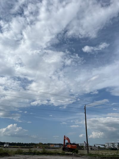 This is a landscape photograph taken in Palmetto, United States. The image depicts a construction site under a cloudy sky with scattered cumulus clouds. In the foreground, there is disturbed earth and some vegetation. Power lines stretch across the frame, supported by a wooden utility pole. In the middle ground, an orange Hitachi excavator is parked on the site. Behind the excavator, there are piles of dirt and debris, along with some newly constructed buildings that appear to be residential. An American flag is visible behind the excavator. In the far background, more buildings are seen, some of which have a coastal or tropical architectural style. A white utility truck is visible on the left side of the image.