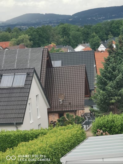 The image shows a residential area in Bad Oeynhausen, Germany, with a view of houses and lush greenery. Several houses with different roof types are visible, including those with dark grey, brown, and orange tiles. Solar panels are installed on one of the roofs, and solar thermal collectors are also present on another. A large evergreen tree stands prominently on the right side of the image. In the background, a hilly landscape covered with trees and a cloudy sky can be seen. The foreground features a well-maintained hedge and part of a translucent roof structure. The presence of the "HUAWEI P30 Pro LEICA QUAD CAMERA" watermark suggests the photo was taken with this device. No people or active interactions are depicted.