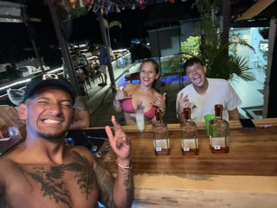 The image, rotated 180 degrees for some reason, depicts three individuals standing behind a bar. The bar has three partially empty liquor bottles and a cell phone sitting on it. A man wearing a white t-shirt and making a "peace" hand gesture is on the left side of the image. In the middle, a woman is wearing a pink bikini top. On the right, a shirtless, muscular man with tattoos on his upper body is making a "hang loose" hand gesture. The background appears to be an outdoor scene at night with a building, a tree, other people, and rows of colorful flags in the distance. Given the context, it is likely this image was taken in Playa del Carmen, Mexico.