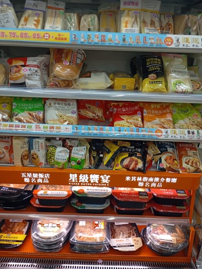 The image shows a refrigerated display shelf in a convenience store, likely in Kaohsiung, Taiwan, filled with pre-packaged food items. The products are arranged on multiple shelves, primarily featuring sandwiches, burgers, rice dishes, and other convenience meals. Many of the packages display pricing in Taiwanese dollars (e.g., 75, 85, 95) and promotional text in Chinese. The overall environment appears to be clean and well-organized, typical of a modern retail setting. No people are visible in the image.