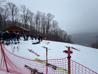 A snowy ski area on a cloudy day. A small building is nestled among bare trees, surrounded by a group of people in ski gear. Skis and poles are scattered across the snow-covered slope, with a red safety net in the foreground. A distant view of a foggy valley is visible beyond the slope.