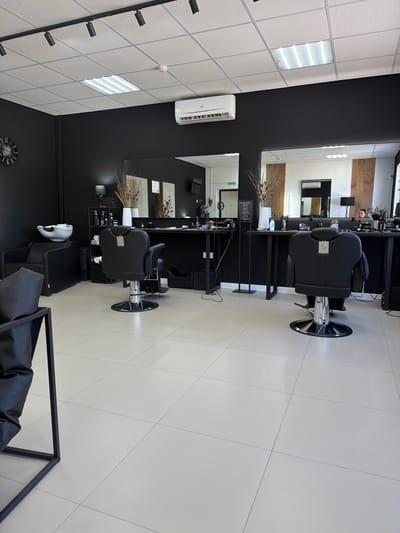 The image depicts the interior of a barbershop in Timashevsk, Russia. The space is modern and minimalist, with black walls and a light-colored tiled floor. Two black hydraulic barber chairs are prominently featured, one in the foreground and another further back. A wash basin with a black seat is visible to the left, next to a black couch. A long black counter runs along the back wall, with mirrors above it. Various barber tools and products are neatly arranged on the counter. The ceiling has a track lighting system and a rectangular fluorescent light fixture. An air conditioning unit is mounted on the wall. A round wall clock with Roman numerals is also visible on the left wall. In the background, a person is partially visible at the counter, appearing to be a client or employee. The overall impression is one of a clean, professional, and contemporary grooming establishment.