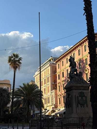 The image depicts a street scene in Sanremo, Italy, looking upwards towards a building with a coral-colored facade and numerous windows. A statue is positioned in front of the building. Palm trees are visible in the foreground and middle ground, framing the view. A blue sky with fluffy white and grey clouds provides a backdrop. Power lines crisscross the sky. The lighting suggests it is daytime, and the perspective implies the photo was taken from street level.