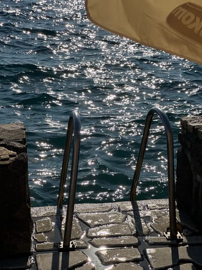 The image shows a view of the Adriatic Sea from a stone-paved platform in Kaluđerac, Montenegro. Sunlight reflects off the water, creating a shimmering effect. A metal ladder with two handrails extends from the platform into the sea. To the right of the ladder, a portion of a large, light-colored umbrella is visible, with a dark logo partially obscured on its surface. The platform is made of irregular-shaped stone slabs with water pooling between them. The overall scene suggests a sunny day at a coastal location.