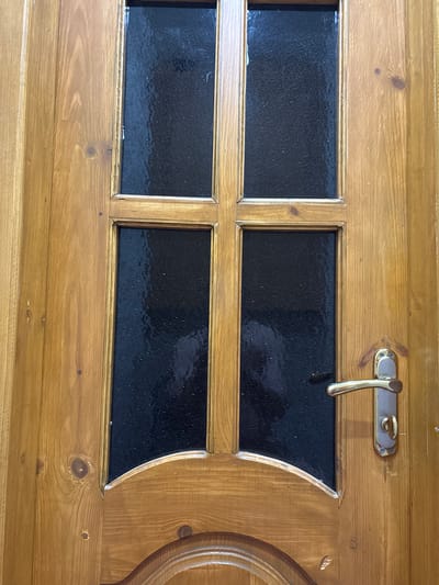 The image shows a wooden door with four rectangular panes of dark, textured glass. A gold-colored door handle is visible on the right side of the door. The door is constructed from light-colored wood planks with visible wood grain and knots. The lower portion of the door shows carved decorative elements. The scene is indoors or in a sheltered area, as indicated by the lighting and the clean appearance of the door. There are no people or animals visible in the image. The location of Novocherkassk, Russia is not discernable from the visual information provided in the image.