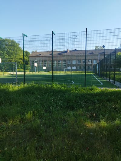 The image depicts an outdoor sports field with artificial turf, enclosed by a chain-link fence with green metal posts. A soccer goal is visible on the left side of the field. In the background, a two-story building with a gray roof and numerous windows is situated behind the sports field. The sky above is clear and blue. The foreground is filled with tall green grass, some of which is illuminated by sunlight. The scene is likely located in Novyi Rozdil, Ukraine, based on the provided context.