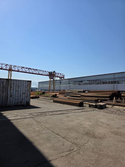The image shows an outdoor industrial scene under a clear blue sky, likely located in Pervomayskiye Prudy, Kazakhstan. In the foreground, a weathered, white shipping container stands to the left, casting a long shadow across a grey asphalt surface. Adjacent to the container are railway tracks. In the middle ground, a large quantity of metal pipes and beams are neatly stacked. A red and yellow gantry crane dominates the mid-ground, positioned over the stacked materials and extending towards a long, silver industrial building with numerous windows in the background. The environment appears to be a storage or processing yard for metal products.