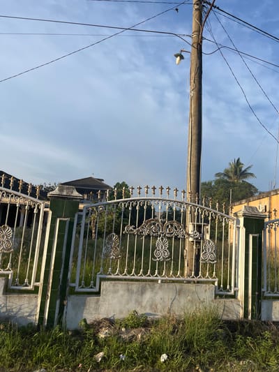 The image depicts a white metal fence with ornate scrolling designs and finials, set against a backdrop of a partly cloudy blue sky. A concrete base supports the fence, with overgrown grass and some litter at its foot. To the right of the fence, a weathered wooden utility pole stands, adorned with a street lamp and numerous electrical wires crisscrossing the frame. Behind the fence, a glimpse of a residential area is visible, featuring a building with a dark tiled roof and trees, including a palm tree. The scene is bathed in the warm light of what appears to be late afternoon or early morning sun. The location is identified as Seunebobaru, Indonesia.