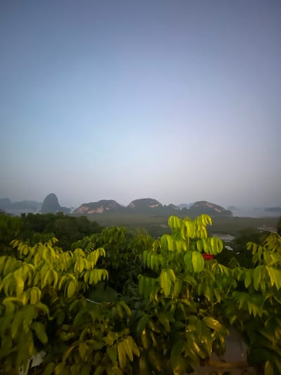 The image depicts a landscape in Khlong Khian, Thailand at dusk. The sky is a gradient of light blue to gray. In the distance, there are mountains partially obscured by haze or mist. Lush green vegetation, including trees and plants with lighter green leaves, fills the foreground and midground on the right side of the frame. The image appears to be rotated 90 degrees counterclockwise.