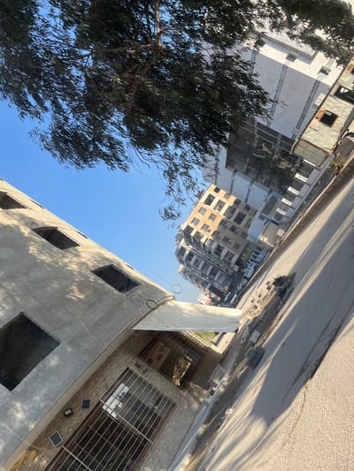 The image depicts a cityscape tilted diagonally, with buildings lining a street above. A tree fills the lower left corner, its leaves dark against the clear blue sky. A concrete building with open window frames dominates the foreground. A building across the street, featuring grilles and a brick facade, is also visible. Debris is scattered along the edge of the street.