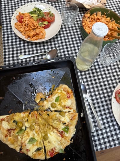 This media file captures an overhead view of a meal set on a black and white checkered tablecloth. The table is laden with food and drinks, suggesting a casual dining experience, likely outdoors given the lighting.
The primary subjects are a partially eaten pizza in a black baking tray and a plate of pasta with tomatoes and avocado. The pizza appears to have toppings of cheese, onions, olives, and basil. The pasta dish includes fresh tomatoes, avocado slices, and microgreens. Two glasses of water are visible, along with a clear bottle of what appears to be lemonade or water, with a label showing "L-117200108 11/2026". There are also wooden tongs in a bowl of pasta, and silverware including spoons and forks.
The scene is set in what appears to be a home environment, possibly a garden or patio, indicated by the dappled sunlight and the relaxed arrangement of the food. The time of day seems to be daytime, likely lunch or early dinner. The mood is one of enjoyment and sharing a meal. The location context suggests this meal is taking place in l'Ametlla del Vallès, Spain.