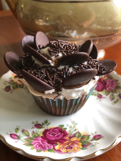 A close-up, overhead shot shows a single cupcake decorated with chocolate shavings and chocolate chips, sitting on a floral-patterned teacup. The teacup has a scalloped edge with gold trim and is adorned with pink and yellow roses. To the left of the cupcake, a portion of a shiny, golden object is visible, possibly a teapot or vase. The cupcake itself is in a blue and white striped wrapper and is topped with white frosting. The overall setting appears to be a dining area or kitchen, suggested by the warm lighting and wooden surface beneath the teacup. The image seems to capture a moment of indulgence, focusing on the decorative details of the dessert. There is no visible text.