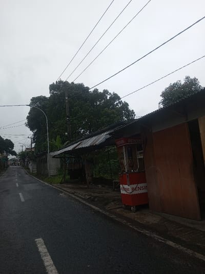 The image displays a damp asphalt road under an overcast sky. On the right side of the road, a roadside stall with a corrugated metal roof is visible. Beneath the roof, a red and white mobile fuel dispenser on wheels is present, labeled with "PERTAMAX" and "BENSIN." This text indicates an Indonesian context. To the immediate right of the dispenser is a closed, light-brown wooden door belonging to an adjacent structure. Overhead, multiple utility lines traverse the scene. A utility pole with attached power lines and a street light pole are positioned further back on the right, amidst dense green foliage and trees that line the road. The asphalt road features white dashed lane markings and recedes into the background, where more indistinct buildings and trees are visible. The ground immediately surrounding the stall is unpaved and uneven. No individuals or specific actions are discernible in the scene.