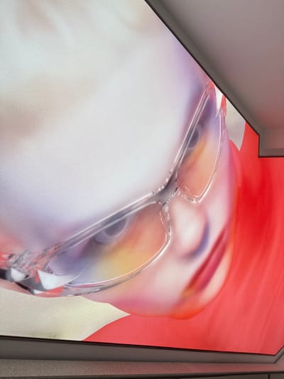 The image presents a low-angle, close-up view of a large, brightly lit digital display or artistic print, integrated into a modern architectural setting in what is likely Shanghai, China. The primary subject on the display is a stylized human face, rendered in soft, ethereal colors, specifically showing the upper part including the eyes and forehead. The face appears to be wearing clear, transparent-framed glasses that reflect ambient light.
The facial features are softly blurred, with gentle gradients of purple, yellow, and pink hues around the eyes and forehead, contrasting with a vibrant red background that transitions into softer tones. The display surface itself exhibits a subtle fabric-like texture, suggesting a backlit tension fabric display or a similar large-format print material.
The display is framed by clean, minimalist architectural elements: a light grey wall and ceiling form an acute angle in the upper left, while a darker grey frame or pillar borders the display on the right. These elements suggest an indoor or sheltered urban environment, possibly a commercial space, gallery, or transit hub within a modern city like Shanghai.
No specific activity or event is taking place; the image captures a static visual display. The lighting emanates from the display itself, creating an artificial glow. There is no visible text. The overall mood is modern, artistic, and serene, focusing on the aesthetic appeal of the displayed face and its interaction with the surrounding architecture.