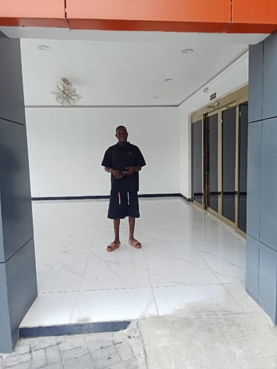 A young man in a black short-sleeved shirt and black shorts is standing in a white-tiled hallway. He is wearing brown sandals. He is holding a black object in his right hand. His left arm is bent at the elbow. His head is tilted slightly to the right, and he is looking toward the right of the image. The hallway is lit by several ceiling lights. There is a large, glass-paned door with a gold frame to the right of the man. The door is closed. There is a gray wall to the left and right of the hallway. The wall on the left is taller than the wall on the right. There is a white ceiling above the hallway. There is a large, metal, decorative fixture with three arms hanging from the ceiling. The coordinates of the location are latitude: 5.472, longitude: 5.817, which is in Ujevwu, Nigeria. The entrance is likely a storefront.