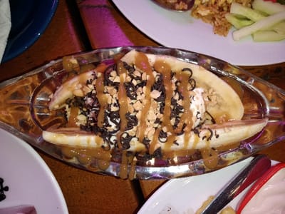 This close-up shot captures a decadent banana split dessert served in a clear, boat-shaped glass dish on a wooden table. The dessert features at least two scoops of white ice cream (likely vanilla) nestled between two halves of a sliced banana. It is generously drizzled with a thick caramel sauce, sprinkled with a substantial amount of small chocolate curls or sprinkles, and topped with chopped peanuts. Some of the caramel sauce has dripped down the sides of the glass dish, indicating it was recently served.

In the background, a white plate holds a portion of what appears to be fried rice, accompanied by slices of cucumber or another green vegetable. Another white plate is partially visible in the bottom-left corner, showing a dark, undefined item. To the bottom-right, part of a plate with a red and white rim is visible, along with what looks like silver tongs or serving utensils.

The scene suggests an indoor dining setting, likely a restaurant or cafe, with warm, low lighting that casts a slight purple or pink hue in the background, possibly from decorative lighting. The focus is clearly on the dessert, which appears rich and appealing. There are no people visible, and no visible text. The overall atmosphere implies an evening meal or dessert occasion.