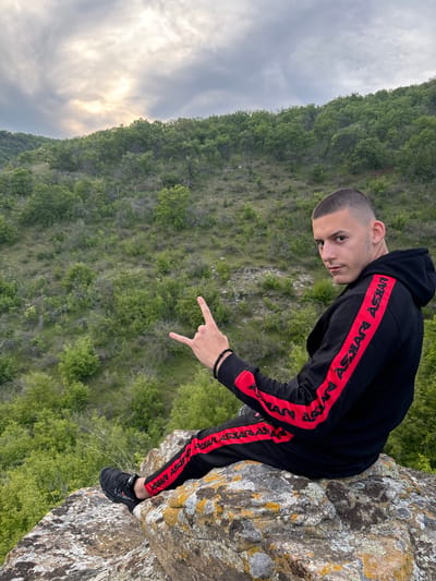 A man in a black hooded tracksuit with red stripes stands on a lichen-covered rock formation, likely in a mountainous region based on the backdrop of dense green trees. He is facing the camera with a slight downward tilt of his head and is making a "rock on" hand gesture. The setting appears to be outdoors, possibly near Goritsa, Bulgaria, given the mention of the city. The sky is overcast, and the overall environment suggests a natural, rugged landscape.