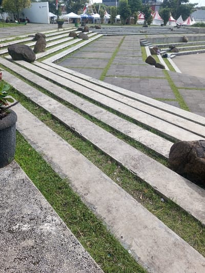 A tiered concrete amphitheater or park space. Steps interspersed with grass lead upward. Scattered boulders punctuate the levels. White tents are visible in the background, suggesting a temporary event or gathering. The structures and greenery suggest a public space designed for relaxation or outdoor performances. A flower pot stands at the edge of the frame.