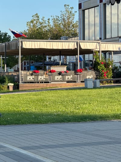 The image shows an outdoor dining area of a restaurant, possibly located in Grushovaya Balka, Russia, based on the presence of the Russian flag. The area is covered by a beige awning supported by metal poles. In front of the awning, a wooden deck is visible, with a railing adorned with banners that display the logo "gogi" and text in Cyrillic. Planter boxes with red flowers are placed along the railing. Beyond the deck, the restaurant building features large, reflective windows. A bush with red roses is prominent on the right side of the image. A patch of vibrant green grass with white clover flowers stretches across the foreground, leading to a paved pathway made of grey tiles. The sky above is clear and blue, with sunlight illuminating the scene.