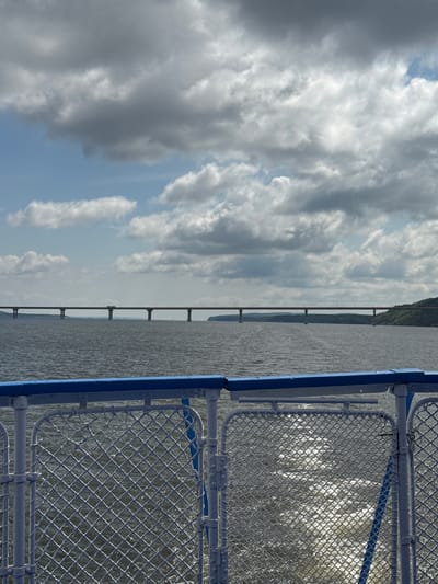 A wide-angle, eye-level shot shows a view from the deck of a boat sailing on a body of water. In the foreground, a blue and white railing with chain-link fencing runs horizontally across the frame. Beyond the railing, the dark, choppy water churns with white foam, indicating the boat's movement. In the midground, a long, wide bridge with numerous support pillars spans the water. The bridge extends across the horizon and appears to be supported by dark, slender pillars. The far background features a distant, wooded shoreline on the right, bathed in soft light. The sky above is filled with a mix of dramatic, heavy grey clouds and lighter, scattered white clouds, with patches of blue sky visible. The scene suggests travel on a river or large lake, possibly on a cloudy day. This view is consistent with the location of the Volga River near Матюшино, Russia, where such a bridge might be found.