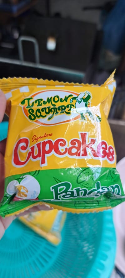 In Mandaluyong City, Philippines, a close-up shot captures a yellow and green packaged product, likely a snack, placed on a turquoise plastic basket. The packaging prominently features the text "Lemon Square" in green script, followed by "Signature Cupcakes" in red script. A small, cartoonish figure dressed in green and white is visible on the right side of the "Lemon Square" logo. Below the "Cupcakes" text, the word "Pandan" is written in green script, accompanied by an illustration of what appears to be an egg or a small pastry. The packaging shows signs of being opened, with a portion of the yellow inner wrapper visible. The background is blurred, suggesting an indoor or partially covered outdoor setting.
