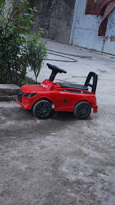 In the city of Pista-mazor, Tajikistan, a red toy car is parked on a dirt and asphalt surface. The car, designed to resemble a sports car, has black wheels and a black steering wheel and seat. To the left of the car, a bush with green leaves and some brown foliage is visible. In the background, a rough concrete wall and a large tree trunk are present. Further in the background, a white door with a closed latch and two oriental rugs hanging on it can be seen. The ground is uneven with scattered debris.