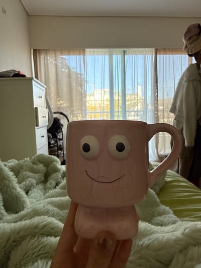 An indoor scene, likely a bedroom, is depicted with a light pink ceramic mug held by a hand in the lower center foreground. The mug has prominent white googly eyes with black pupils, a simple black curved smile, and a rounded handle on the right; its surface exhibits fine crackling. Below the mug, a light green, textured fuzzy blanket covers a bed. To the left, a white chest of drawers with dark pull handles is visible, behind which a dark, indistinct object is partially seen. To the right, a person is partially visible, wearing a light-colored, possibly white, fluffy garment and a light pink or beige hat, facing away from the camera. The background features a large window or glass door covered by translucent, light-brown sheer curtains. Beyond the curtains, green and yellow-leafed foliage is visible, along with a light-colored building under a blue sky. The scene is brightly illuminated by natural light from the window. The specific location within Kalyvia Thorikou Municipal Unit, Greece, is not discernible from the visual elements present.