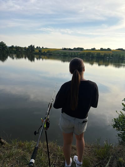 An outdoor, medium shot captures a person from behind, fishing by a tranquil body of water. The individual, with long, dark hair tied in a ponytail, wears a black t-shirt and light-colored denim shorts. A fishing rod is set up on a stand to the left of the person, its tip extending towards the water. The background reveals a calm lake reflecting the sky and a distant shoreline with trees and a field of yellow flowers, characteristic of the rural landscape of Tătărăști, Moldova. The sky above is partly cloudy, with soft daylight illuminating the scene. The foreground consists of grassy, dry terrain with some twigs. The overall impression is one of peaceful solitude and outdoor leisure.