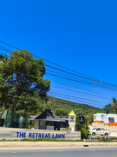 This is a wide-angle, eye-level outdoor shot of a street-level view in Baan Thong Po, Thailand, on a clear, sunny day. The foreground features a light-colored concrete barrier with the blue, three-dimensional letters "THE RETREAT LAWN" prominently displayed. To the right of the barrier, a white pickup truck is parked near a modern building complex with white walls and orange accents.
Behind the barrier, a small, dark-roofed pavilion-like structure stands, with a white wall visible behind it. A short distance to the right of this structure, a yellow tiered monument or decoration is situated, topped with a golden spire. A black circular sign with white text hangs from a pole nearby, reading "RETREAT LAWN CAFE". A large, leafy green tree dominates the left side of the frame, with numerous power lines crisscrossing in front of it and extending across the upper portion of the image. In the background, a lush green hillside rises, dotted with palm trees, and the bright blue sky fills the upper half of the image. The scene appears to be a commercial or hospitality area along a road.