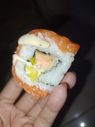 A close-up shot captures a hand holding a piece of sushi, likely in a dimly lit indoor setting, potentially a home or restaurant, with a dark, out-of-focus background. The sushi roll, with its vibrant orange salmon on the outside, is topped with a creamy white sauce and appears to contain white rice, green elements, and possibly another filling. The hand, belonging to a person with neatly manicured nails, gently supports the sushi, showcasing the texture of the skin and the slight sheen from the sauce. The image is tightly cropped on the sushi and hand, suggesting a focus on the food item itself. The overall impression is one of enjoying a meal, with the dark background emphasizing the subject. There is no information available to determine the specific location within Pasir Gadung, Indonesia.