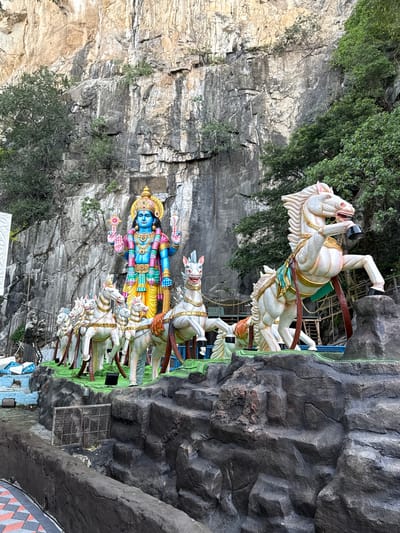 This image captures a grand, colorful statue display set against a natural cliff face. The central figure is a large, blue-skinned deity, likely Lord Vishnu or Lord Krishna, depicted with four arms, holding a chakra (discus) and a conch shell among other attributes. The deity is richly adorned with a golden crown, intricate jewelry, and vibrant clothing including a yellow dhoti and a blue and green upper garment.

The deity stands on an elaborate chariot pulled by seven white horses. The horses are sculpted in dynamic, rearing poses, conveying motion, and are decorated with ornate saddles and bridles. The entire chariot and horse ensemble rests on a green-colored platform.

The backdrop is a massive, rugged limestone cliff, showcasing natural strata and crevices. Green foliage and trees are visible on the cliff face and atop it. To the right, artificial dark grey rock formations frame the scene, mimicking the natural environment. The bright lighting indicates daylight, and the sky appears clear. This impressive religious sculpture, located in Selayang Municipal Council, Malaysia, creates a striking visual of mythology intertwined with a natural landscape. No text is visible in the image.