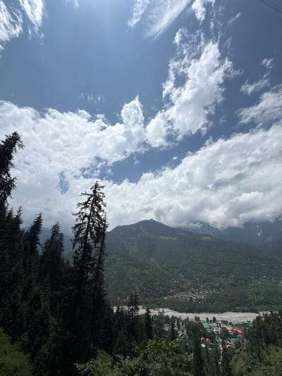 The image is an outdoor, high-angle shot of a mountainous landscape under a partly cloudy sky. In the foreground, a dense forest of dark green coniferous trees dominates the left side of the frame. These trees are tall and irregular in shape, with some extending towards the top of the image.
Further into the distance, the terrain slopes downwards, revealing a valley with a village nestled within it. The village appears to be located near a river or dry riverbed filled with stones. The buildings in the village are small and colorful, with many featuring red roofs. The valley is surrounded by verdant, tree-covered mountains. The sky is a bright blue, with large, white, fluffy clouds scattered across it, particularly in the upper half of the image.
Based on the provided context, this scene is located in the city of Bahang, India.