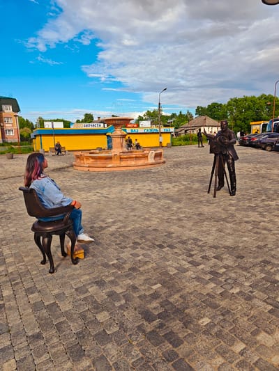 The image captures a public square in Faliukovo, Russia. The square is paved with patterned brickwork. A brown, multi-tiered fountain is visible. A woman with dyed pink and blue hair sits on a dark-colored chair, elevated by a wooden block, facing the fountain. Further away, a bronze statue of a man with a camera on a tripod faces the woman. People are seated on benches around the fountain. In the background, a yellow building with blue trim, reading "Avto Zapchasti Gazel UAZ" in Russian, can be seen, alongside a two-story building and parked cars. The sky is partially cloudy with patches of blue. Streetlights are positioned around the square.