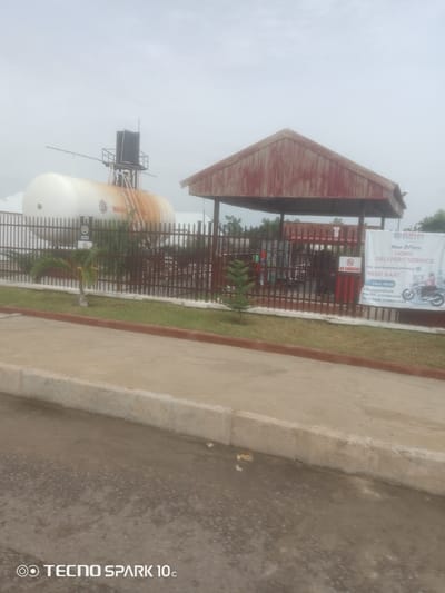 This media file depicts an outdoor scene at a gas station in Zanzi, Nigeria. The primary subjects are large storage tanks, likely for gas, with one prominent white tank featuring the "BRIM ENERGY" logo. Adjacent to the tank is a covered refueling area with multiple gas cylinders visible behind a metal fence. A "NO SMOKING" sign is posted, indicating safety regulations.

A banner on the right side of the refueling area advertises "HOME DELIVERY SERVICE" for gas, providing contact numbers for different locations within Nigeria. The banner also features an illustration of a person on a motorcycle, suggesting delivery logistics.

The setting appears to be a functional gas station with paved ground and a concrete curb in the foreground. A small palm tree and some green grass are visible in front of the fence, adding a touch of landscaping to the industrial environment. The sky is overcast, suggesting a cloudy or hazy day, and the time of day is indeterminate. There are no people clearly visible in the image, but the presence of refueling equipment and delivery service promotion implies ongoing operations. The overall impression is one of a commercial establishment providing essential services. The bottom left corner of the image contains a watermark "TECNO SPARK 10c".