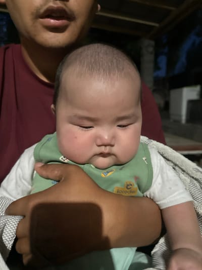 An adult with dark hair and facial hair, wearing a maroon shirt, holds a baby. The baby has light skin, a round face with rosy cheeks, and dark hair. The baby is wearing a green bib with a yellow pattern and a white shirt underneath. The baby is being held close to the adult and appears to be looking down. A grey and white patterned blanket is draped over the adult's arm and the baby. The background is dark and appears to be an outdoor setting, possibly a porch or patio, with some blurry structures and foliage visible. The location is specified as Тайгараев, Kyrgyzstan.