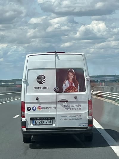 The media file shows the rear of a white van, likely a Volkswagen Crafter, with several details visible. On the back doors, there are company logos and text. The main logo appears to be for "bunexim" with "SINCE 1994" written within a circular emblem. Social media icons for Facebook, Instagram, and TikTok are also present, along with the text "Bunexim" again.
To the right of the logos, there is contact information: phone numbers "0259 427 596" and "0359 427 410", a website "www.bunexim.ro", and an address "Oradea, Ardealutul 14". A license plate is clearly visible, reading "B 210 VIM". There is also a smaller text which appears to be a phone number "021-314 35 54".
A prominent feature on the left rear door is a large sticker or decal featuring a young woman with long brown hair. She is wearing a traditional-looking garment with red and white embroidery and a flower crown, and she is holding a bouquet of what appear to be red flowers or fruits.
The van is on a road, possibly a highway, with a bridge structure in the background and a railing along the side. The sky is cloudy, suggesting a daytime scene. The overall impression is of a commercial vehicle advertising its services and its brand. The presence of the woman in traditional attire might suggest a connection to cultural events, tourism, or a specific product/service related to Romanian traditions. The location context is Arad, Romania.