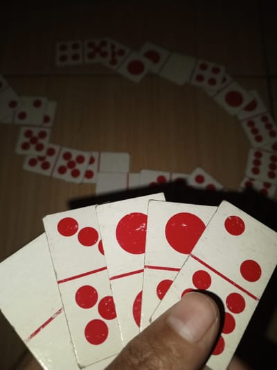 A close-up, low-light photograph from Kpein, Liberia, depicts a game of dominoes in progress. In the foreground, a human hand holds five white rectangular domino tiles, each featuring red pips. The visible faces of these held tiles include combinations such as two pips over four, one pip over three, one pip over three, and two pips over two, moving from left to right. In the background, numerous other white domino tiles with red pips are arranged on a light-brown wooden surface. These tiles form a long, winding, interconnected chain, characteristic of active gameplay. The scene is dimly lit, casting soft shadows and emphasizing the red pips against the white tiles and the wooden surface.
