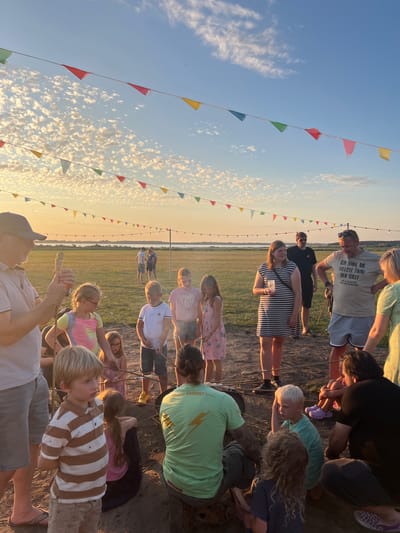 In the city of Am Salzhaff, Germany, a group of people, including adults and children, are gathered outdoors during a sunset. They appear to be participating in a bonfire or a similar outdoor activity, as indicated by the sticks held by several individuals, some of which are positioned near a fire pit. Colorful triangular flags are strung overhead, adding a festive atmosphere. The background features a flat, grassy field stretching towards a body of water and a horizon under a sky filled with scattered clouds and the warm hues of sunset. The overall scene suggests a casual social gathering.