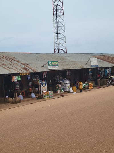 The image captures a street scene in Tangu, Nigeria, featuring a row of small shops with corrugated metal roofs. A tall, lattice-style radio or cell tower dominates the background. The foreground shows a dusty, reddish-brown road. The shops display a variety of goods, including packaged food items, drinks, and what appear to be agricultural supplies like sacks of fertilizer or grain. Several people are visible, some near the shops, possibly vendors or customers. One person stands by the side of the road in front of a shop, while others are seated inside or near the entrances. The overall environment appears to be a bustling marketplace or commercial area.