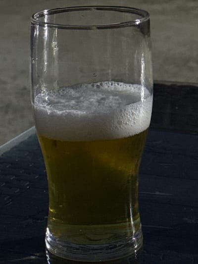 This is a close-up, side-view, horizontal shot of a glass containing a golden-yellow liquid with a significant amount of white foam. The glass appears to be clear and has a rounded rim. The foam occupies the left side of the glass, with the liquid filling the right side. The scene is dimly lit, with the light source likely coming from the left, casting highlights on the rim and the foam. The background is dark and out of focus, with some vertical lines visible, suggesting a textured surface or possibly blinds. The focus is entirely on the glass and its contents. There are no people or discernible emotions visible in the image. The overall impression is that of a close-up detail of a beverage, possibly beer, in a glass. The location context of Khazar Raion, Azerbaijan, does not provide any specific visual cues within the image itself. No text is visible.