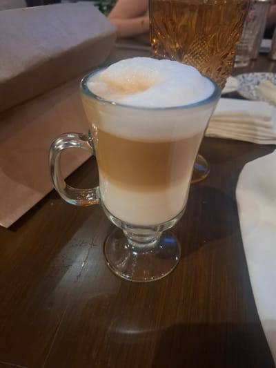 A latte with a thick layer of foam is on a dark wooden table. A decorative glass pitcher sits beside it. Jesús del Monte, Mexico is not discernible in this image.