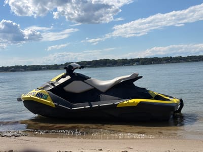 A yellow and black jet ski is partially submerged in the water, resting on a sandy beach. The jet ski is positioned at an angle, with its front end facing towards the left of the frame. The water is calm and appears to be shallow near the shore. In the background, a line of trees and houses stretches across the horizon, indicating a residential area. The sky is a clear blue with scattered white clouds. The location is Hampton Bays, United States of America. There are no people visible in the image.
